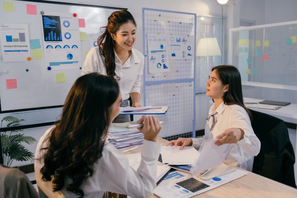 Three asian businesswomen are analyzing charts and documents, collaborating on a project in a modern office environment, showcasing teamwork and data analysis